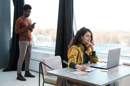 Young businesswoman working on laptop at the table with man standing behind her and using mobile...