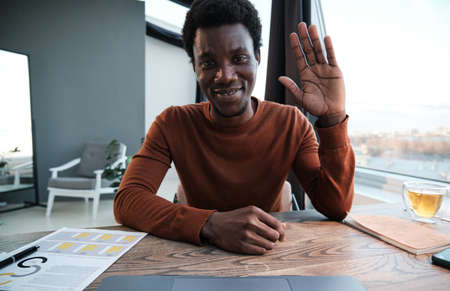 Portrait of African businessman smiling at camera and waving his hand while sitting at office