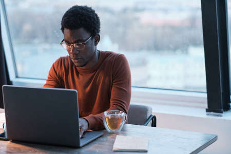African young businessman sitting at the table and typing on laptop he working till late eveningの写真素材