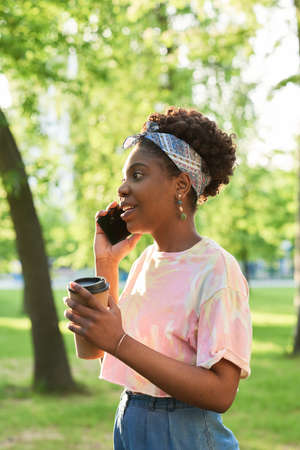 African young woman drinking coffee and talking on mobile phone while standing outdoorsの写真素材
