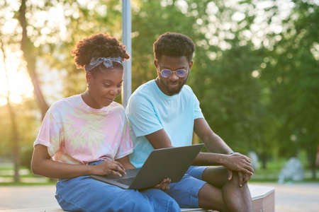 Two friends sitting on the bench nd using laptop together in the parkの写真素材