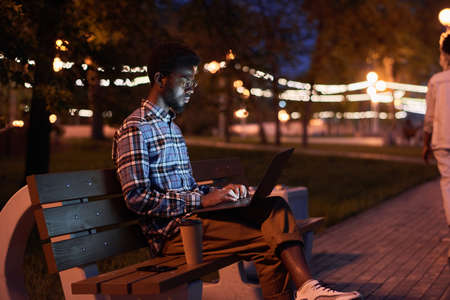 African young man sitting on bench and using laptop in the park in the eveningの写真素材