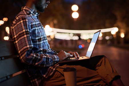 Young man sitting on the bench and working online on laptop in the park in the eveningの写真素材