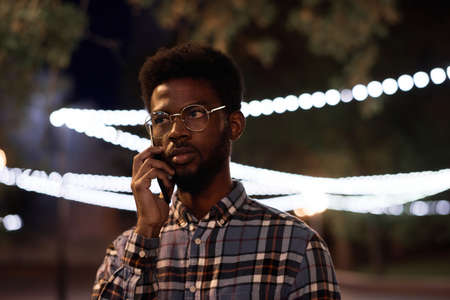African young man in eyeglasses talking on mobile phone while standing in the city in the eveningの写真素材