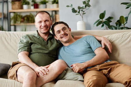 Portrait of happy male gay couple smiling at camera while sitting on sofa in the roomの写真素材