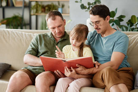 Gay parents sitting on sofa and reading a book to their little daughter in the roomの写真素材