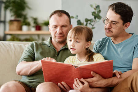 Gay family teaching little girl to read a book while sitting on sofa in the roomの写真素材