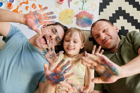 High angle view of happy gay family lying on the floor with their daughter showing their painted hands and smiling they painting at homeの写真素材