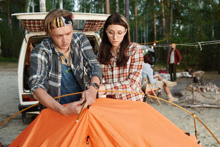 Two hikers setting up the tent together during camping with other people outdoors
