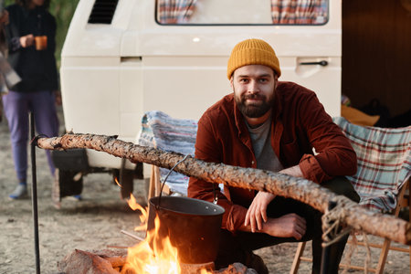 Portrait of young bearded man looking at camera while cooking food on a fire during camping in the forestの写真素材
