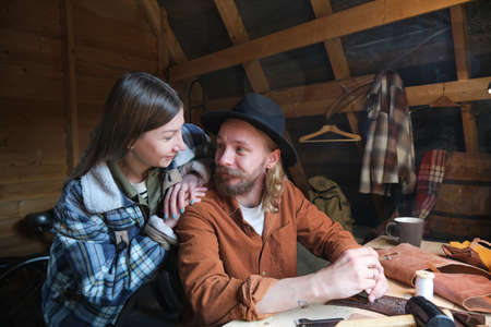 Young man sitting at the table and working with leather with young woman interrupting and talking to...