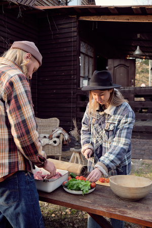 Young woman cutting vegetables for salad at the table with man cutting meat for barbecue, they standing outdoorsの写真素材