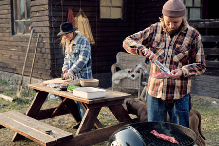 Young man frying meat on barbecue with woman cutting vegetables for salad in the background, they...