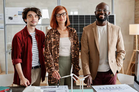 Portrait of successful group of architects smiling at camera while standing at the table with...