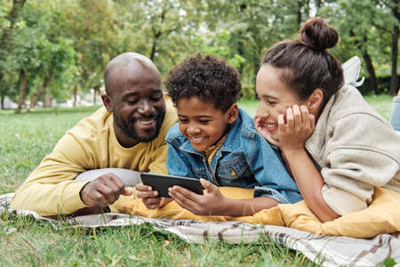 African happy family lying on the grass in the park watching photos on mobile phone and laughingの写真素材