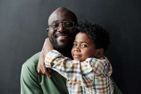 Portrait of African little son embracing his happy father and they smiling at camera isolated on black backgroundの写真素材