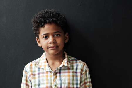 Portrait of African little boy with curly hair looking at camera while standing against the black backgroundの写真素材
