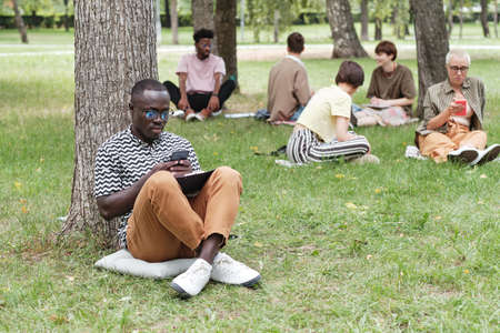 African young businessman sitting on the grass outdoors and using his mobile phone at work with...