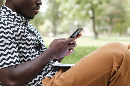 Close-up of African man typing a message on his mobile phone while sitting in the park outdoorsの写真素材
