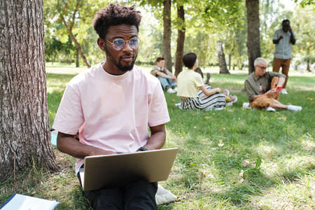 Portrait of African young man in eyeglasses sitting on the grass and typing on laptop, he working or...