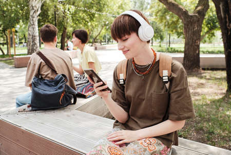 Young woman in wireless headphones and with backpack behind her back sitting on the bench in the park and reading a messageの写真素材