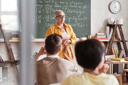Maths teacher standing at blackboard and explaining material to students in the classroomの写真素材
