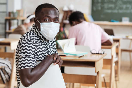 Portrait of African student in mask looking at camera while sitting at desk during lesson at schoolの写真素材