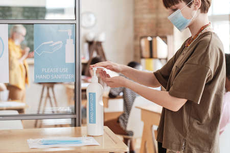 Young woman in mask washing her hands with hand sanitizer before entering to the classroom at schoolの写真素材