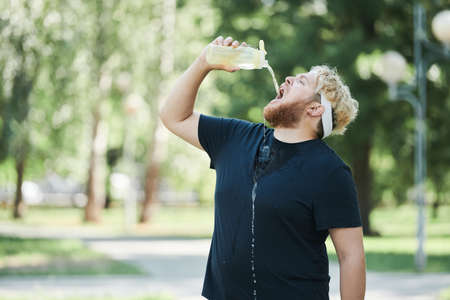 Overweight man drinking water from the bottle during his sport training outdoors in the parkの写真素材