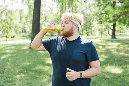 Overweight bearded man drinking fresh juice from bottle after sports training outdoorsの写真素材