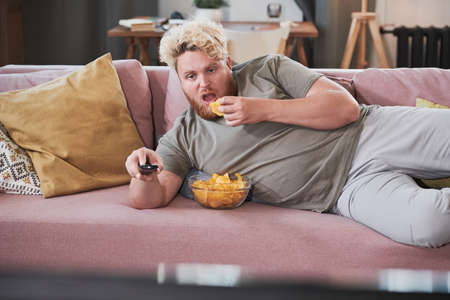 Lazy overweight man lying on the sofa eating chips and watching TV at homeの写真素材