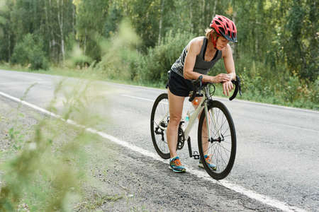 Athlete in helmet checking her fitness bracelet before riding on bike on the country roadの写真素材