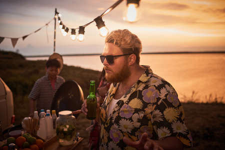Young man in sunglasses holding bottle of beer and dancing with his friends at beach party outdoorsの写真素材