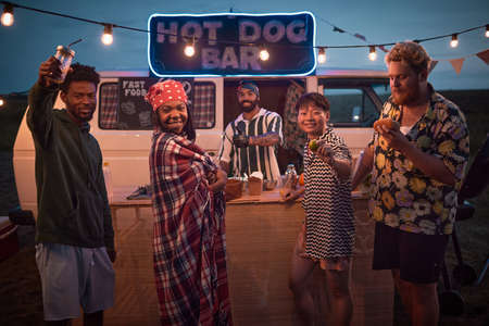 Portrait of young people posing at camera while having fun with food and drinks at beach party with...