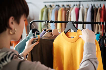 Rear view of young woman looking at clothes on rack in her hands and choosing a new style for herself in the clothes storeの写真素材