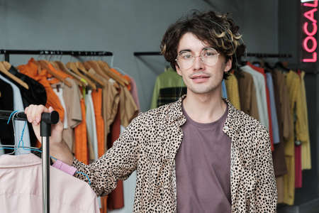 Portrait of young fashion designer in eyeglasses looking at camera while standing in his clothes storeの写真素材