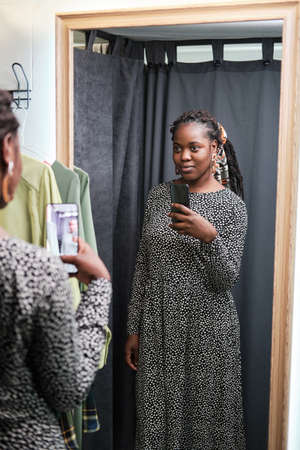 African young woman in new dress looking at the mirror and making selfie on her mobile phone while standing in the fitting roomの写真素材