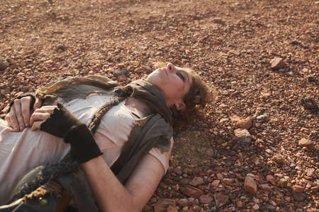 Young woman with dirty face and torn clothes lying on the ground with her eyes closed in the desert