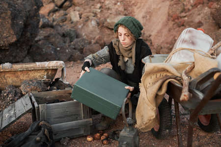 Young homeless woman in torn clothes taking the box out among her things, she living outdoors