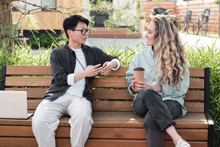 Horizontal medium long portrait shot of two multi-ethnic young women wearing casual clothes sitting together on bench outdoors drinking coffee and chatting during breakの写真素材