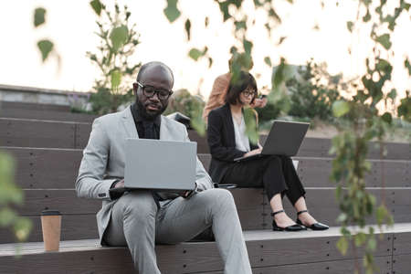 Horizontal shot of modern man and women sitting outdoors on stair benches working on laptops during...