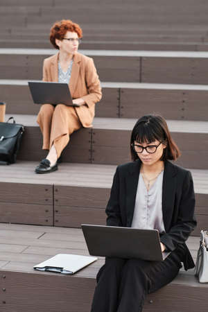 Vertical shot of two female office workers sitting on wooden stair benches outdoors on summer day...