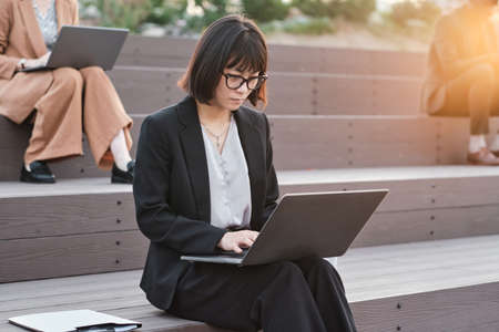 Horizontal medium portrait shot of young Asian woman sitting on wooden bench and working on wireless laptop outdoorsの写真素材