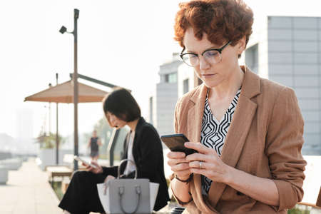 Horizontal medium portrait shot of mature woman with short red hair sitting on bench outdoors...