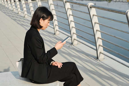 Horizontal shot of serious young Asian woman wearing fashionable elegant clothes sitting outdoors checking her workflow plan on smartphone, copy spaceの写真素材