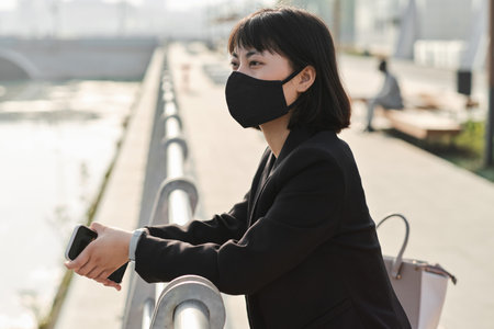 Horizontal medium portrait of modern Asian woman wearing protective mask on face standing on embankment looking at river and thinking of somethingの写真素材