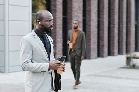 Horizontal medium shot of modern African American man wearing earbuds standing in business district...