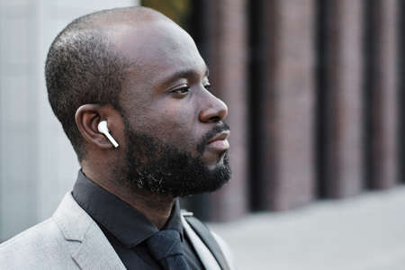 Horizontal side view close-up portrait of serious African American man wearing earbuds standing outdoors listening to music thinking about somethingの写真素材