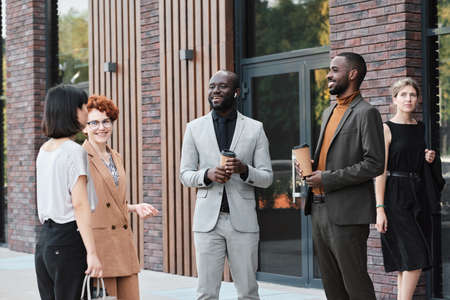 Medium long shot of multi-ethnic co-workers standing outdoors drinking coffee and chatting about funny things during breakの写真素材