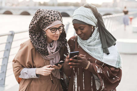 Two friends in muslim clothing discussing photos online on mobile phone during their walk in the cityの写真素材